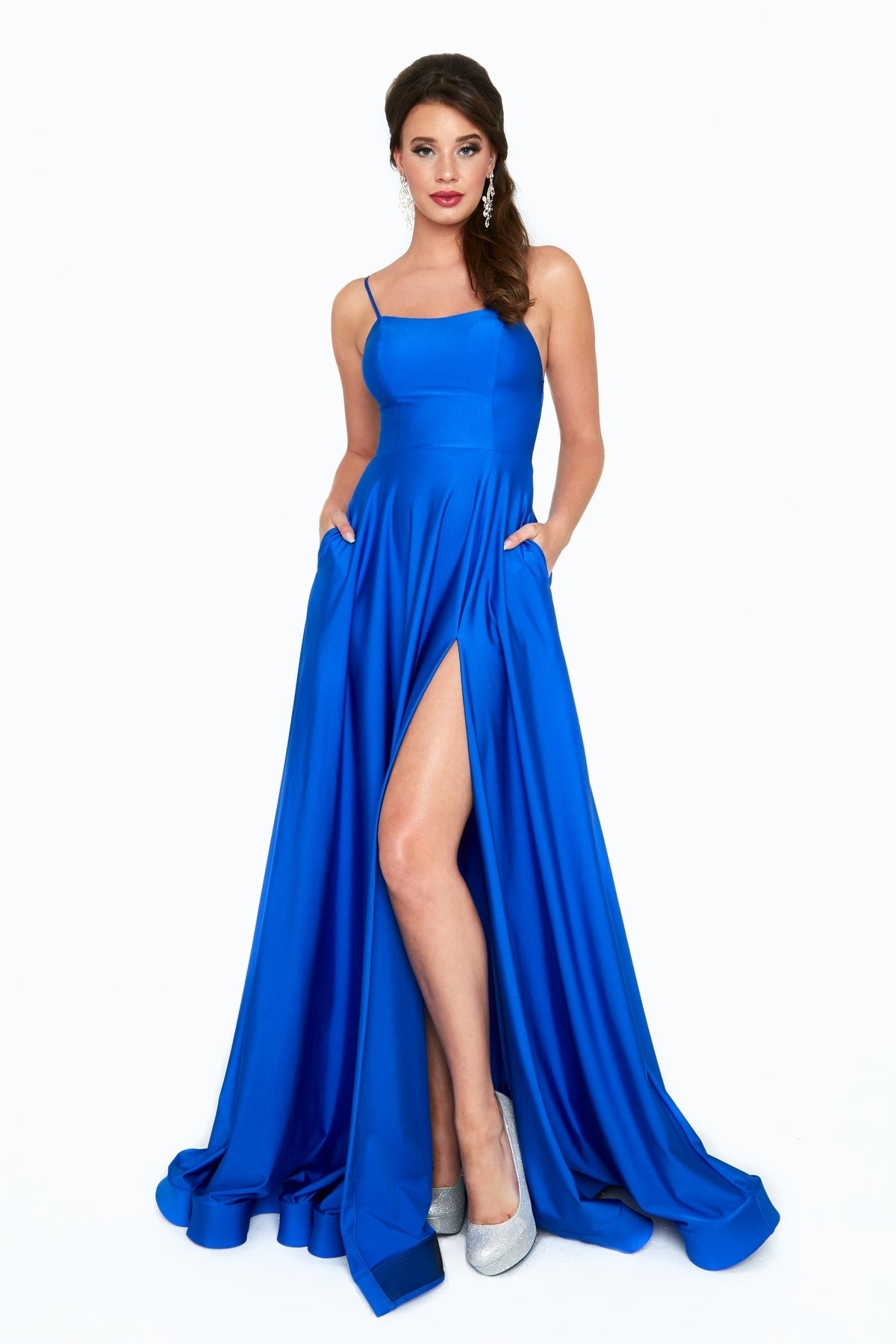 A woman stands facing forward, hands in the pockets of a bright blue Long A-Line Prom Dress with Pockets: Atria 6521H, featuring a thigh-high slit and paired with silver high-heeled shoes.